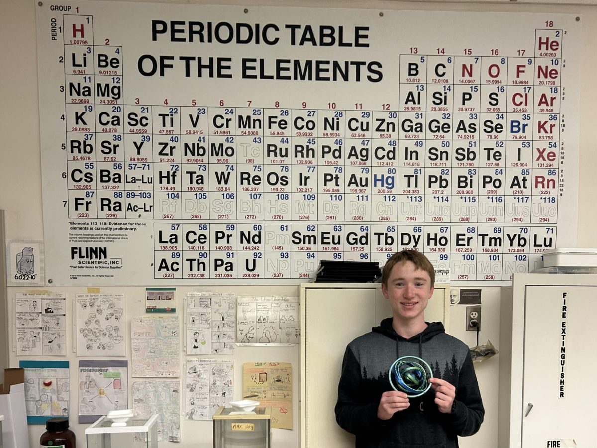 Whitman stands in the his AP Chemistry classroom, with the periodic table behind him. He is holding a self-printed 3D model of an atom. 