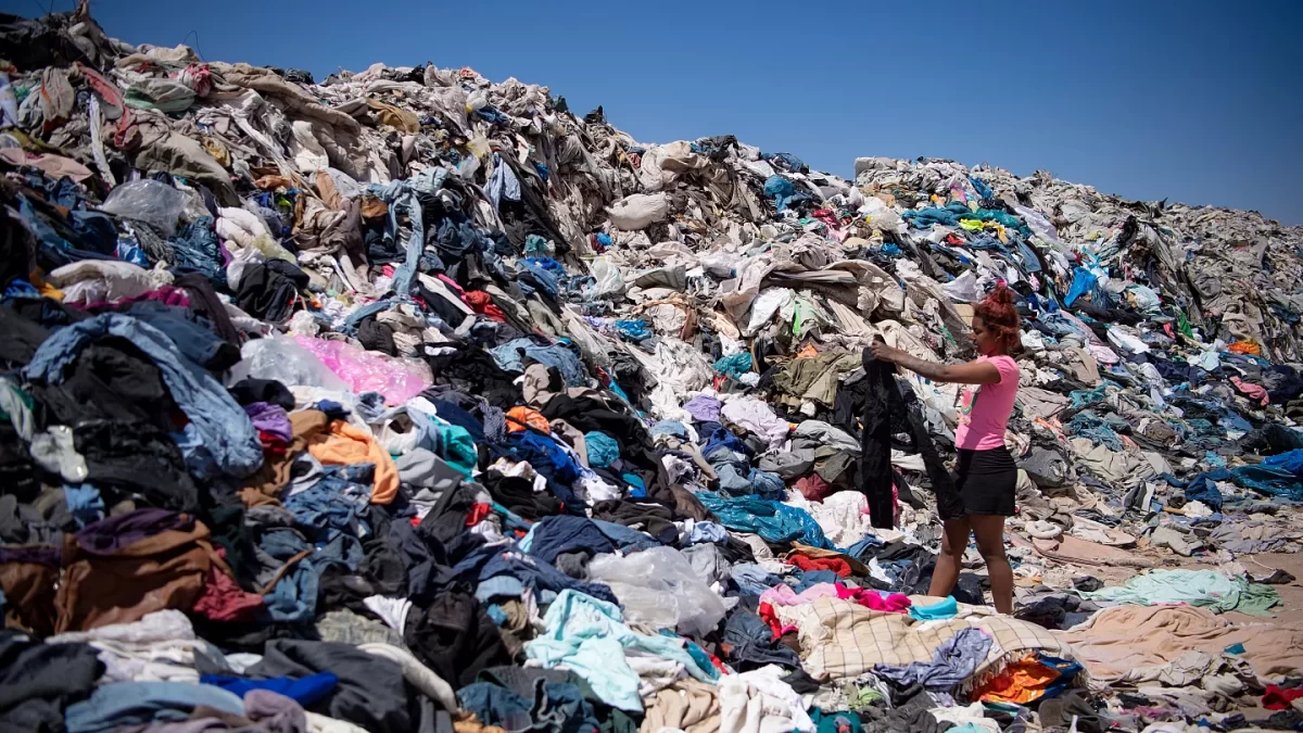 A woman holds up a pair of black jeans amidst a growing pile of clothes in a landfill.