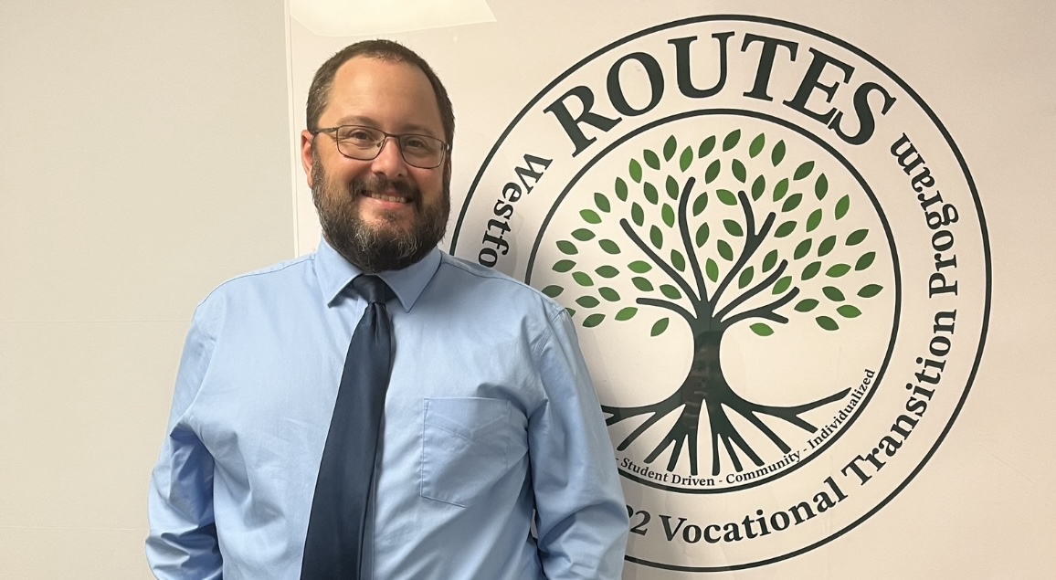 Todd Foster outside his office standing in front of the Routes Program poster.